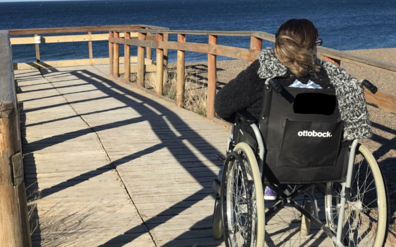 Persona en silla de ruedas recorriendo una pasarela de madera accesible frente al mar en un mirador costero.